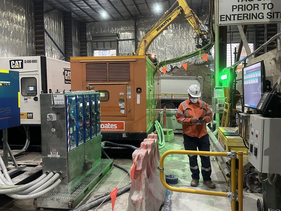 Worker checking equipment on a handheld beside generators and plant inside a site tool shed